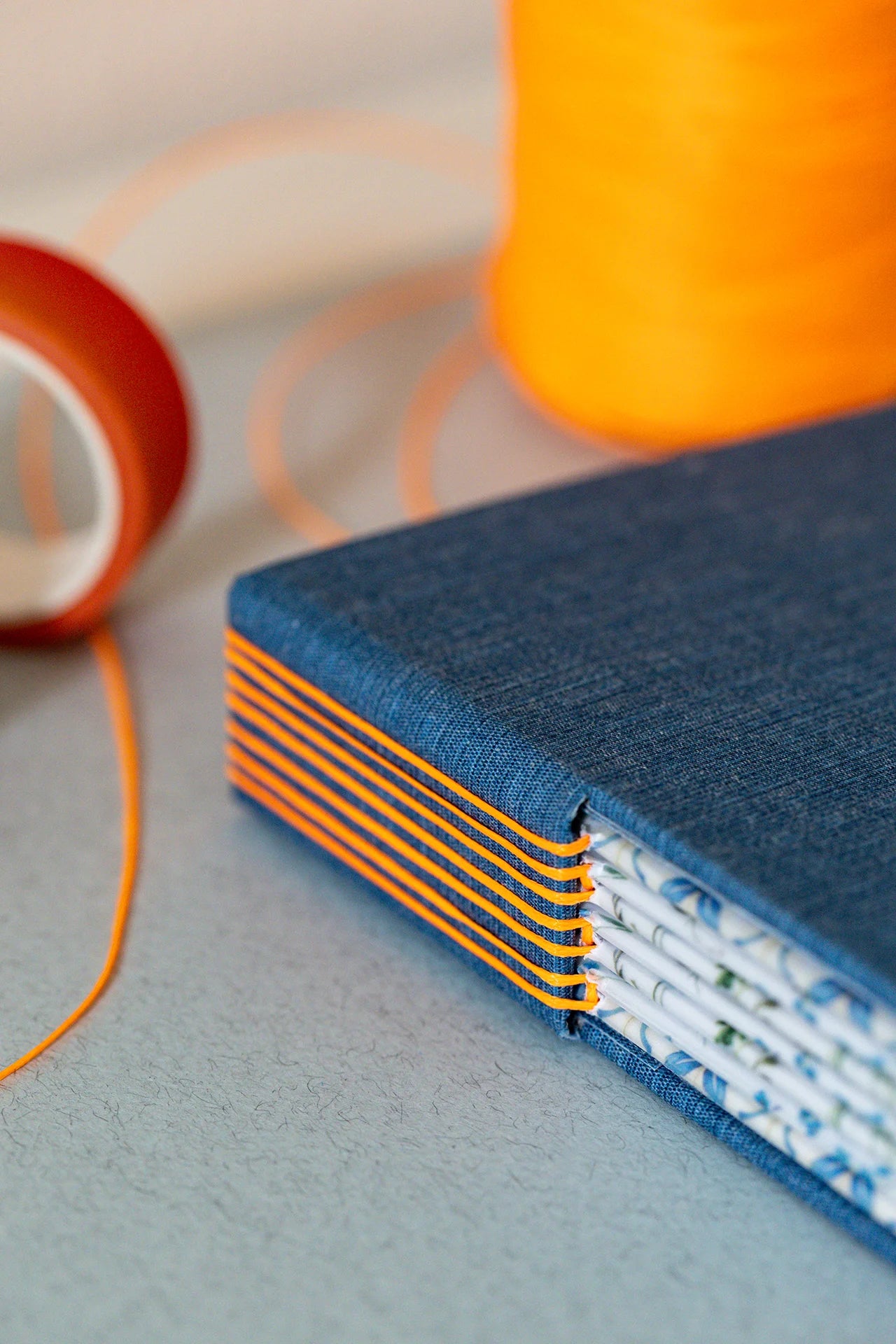Dettaglio di un taccuino sartoriale con foro  sul dorso che lascia intravedere le carte interne decorate con motivi floreali e geometrici, il dorso è rilegato con filo arancione fluo che contrasta sulla tela blu.