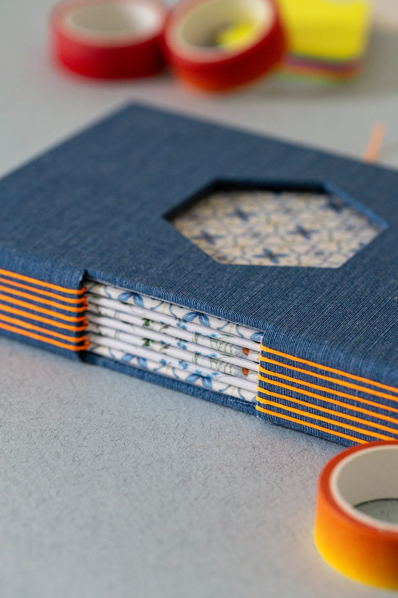 Dettaglio di un taccuino sartoriale con foro esagonale in copertina e finestra sul dorso che lascia intravedere le carte interne decorate con motivi floreali e geometrici, il dorso è decorato dal filo arancione fluo che contrasta sulla tela blu.