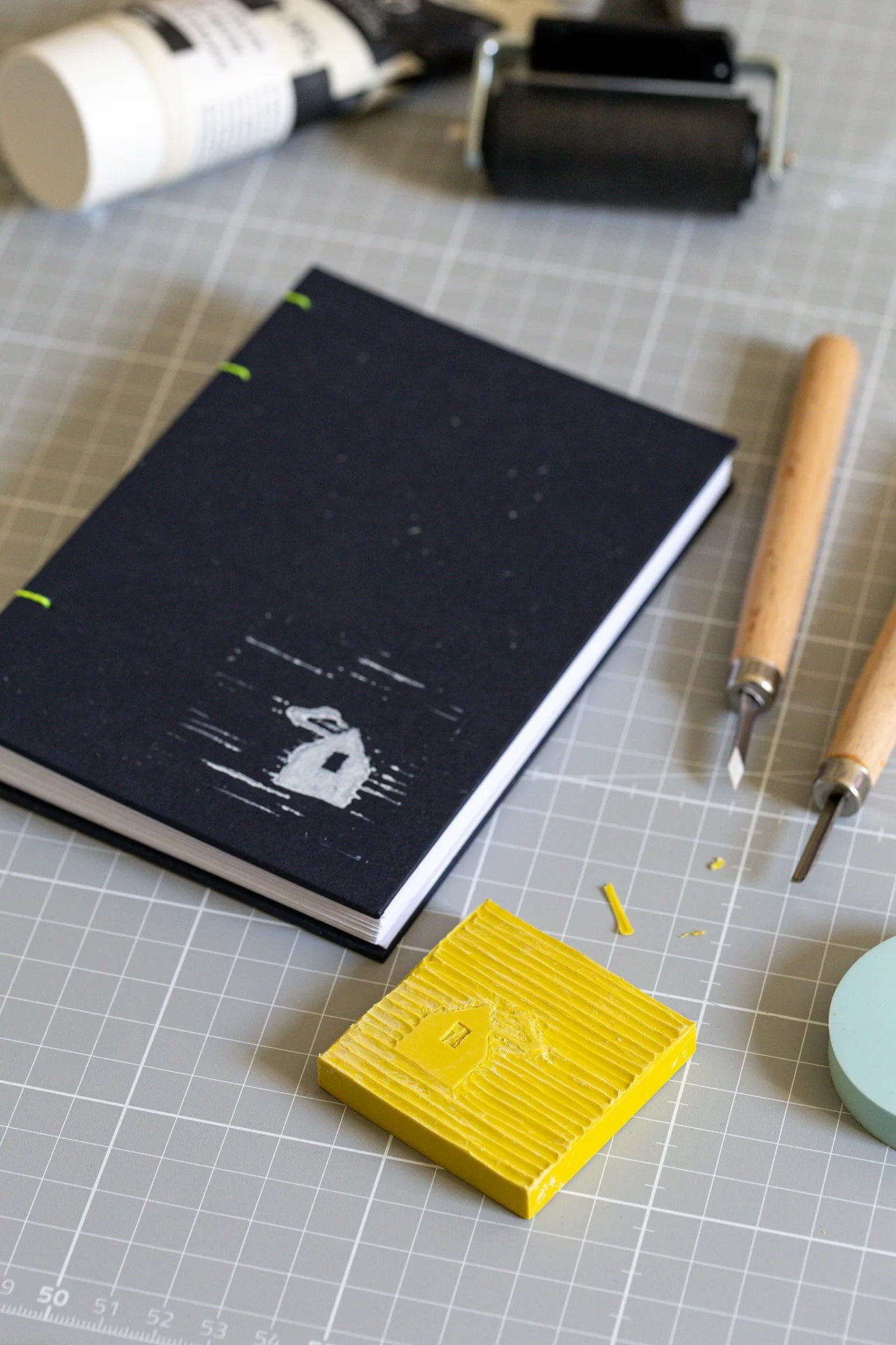 Taccuino A6 con copertina blu scuro in carta da upcycling con legatura artigianale copta in filo fluo, stampa bianca in copertina di una casetta con tecnica linocut e materiali di stampa: rullo, sgorbie, linoleum, inchiostro.