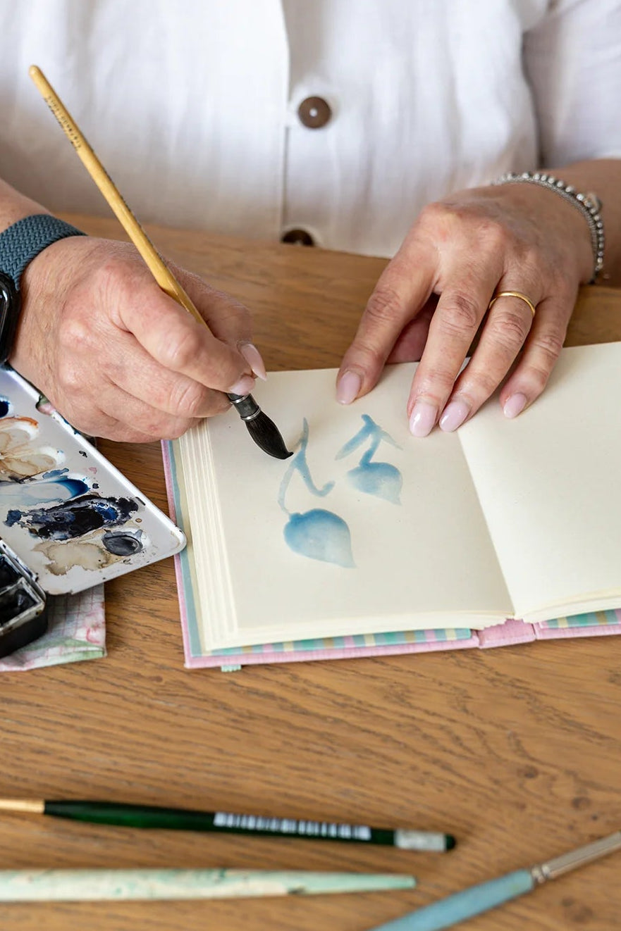 Quaderno d'arte Saimai pezzo unico sartoriale fotografato in atelier di pittura, mani che dipingono un acquerello su quaderno con carta pregiata, apertura piatta a 180 gradi senza ingombri.
