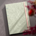 Taccuino rilegato a mano con copertina in carta sostenibile Crush Kiwi di Favini con rilegatura copta in filo verde acqua e elastico di chiusura rosa, foto con fuorifuoco di fiori e ombre naturali.