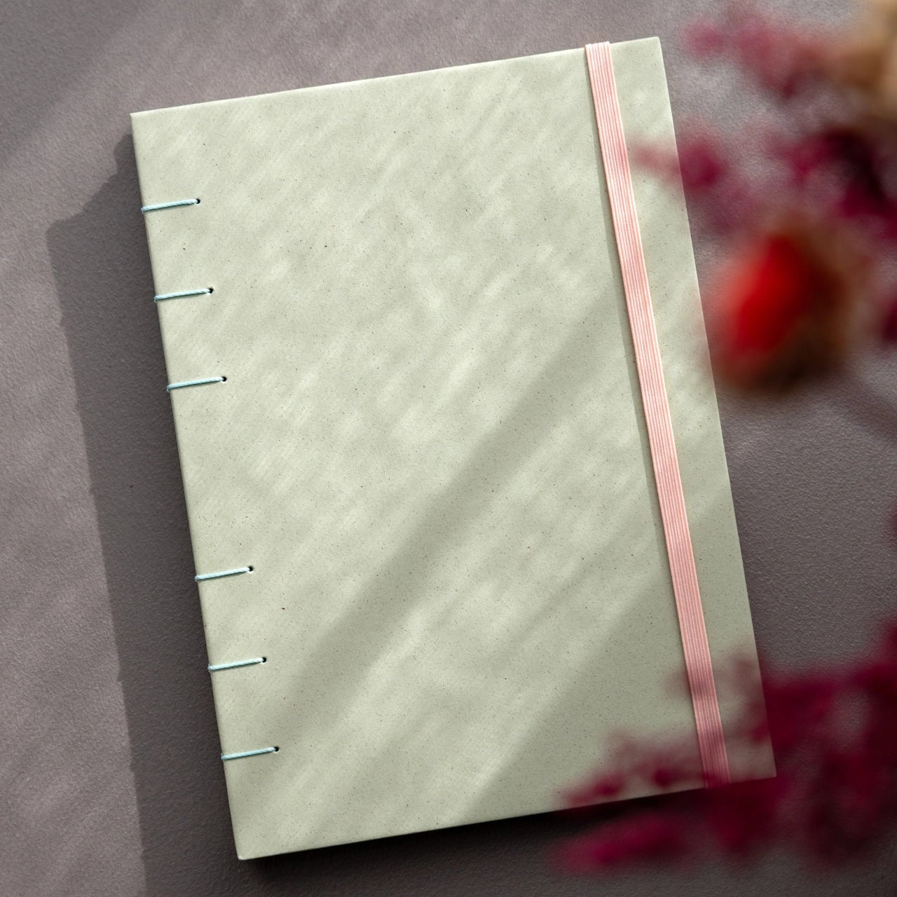 Taccuino rilegato a mano con copertina in carta sostenibile Crush Kiwi di Favini con rilegatura copta in filo verde acqua e elastico di chiusura rosa, foto con fuorifuoco di fiori e ombre naturali.